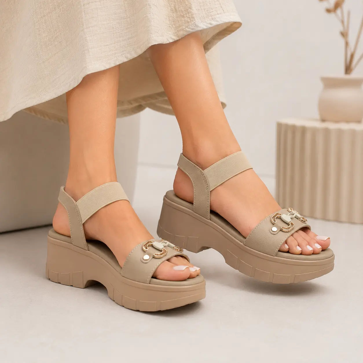 A pair of women's platform sandals in a light taupe-beige color, worn by a model. The sandals feature a chunky sole, a wide adjustable ankle strap, and a decorative metal buckle on the toe strap.