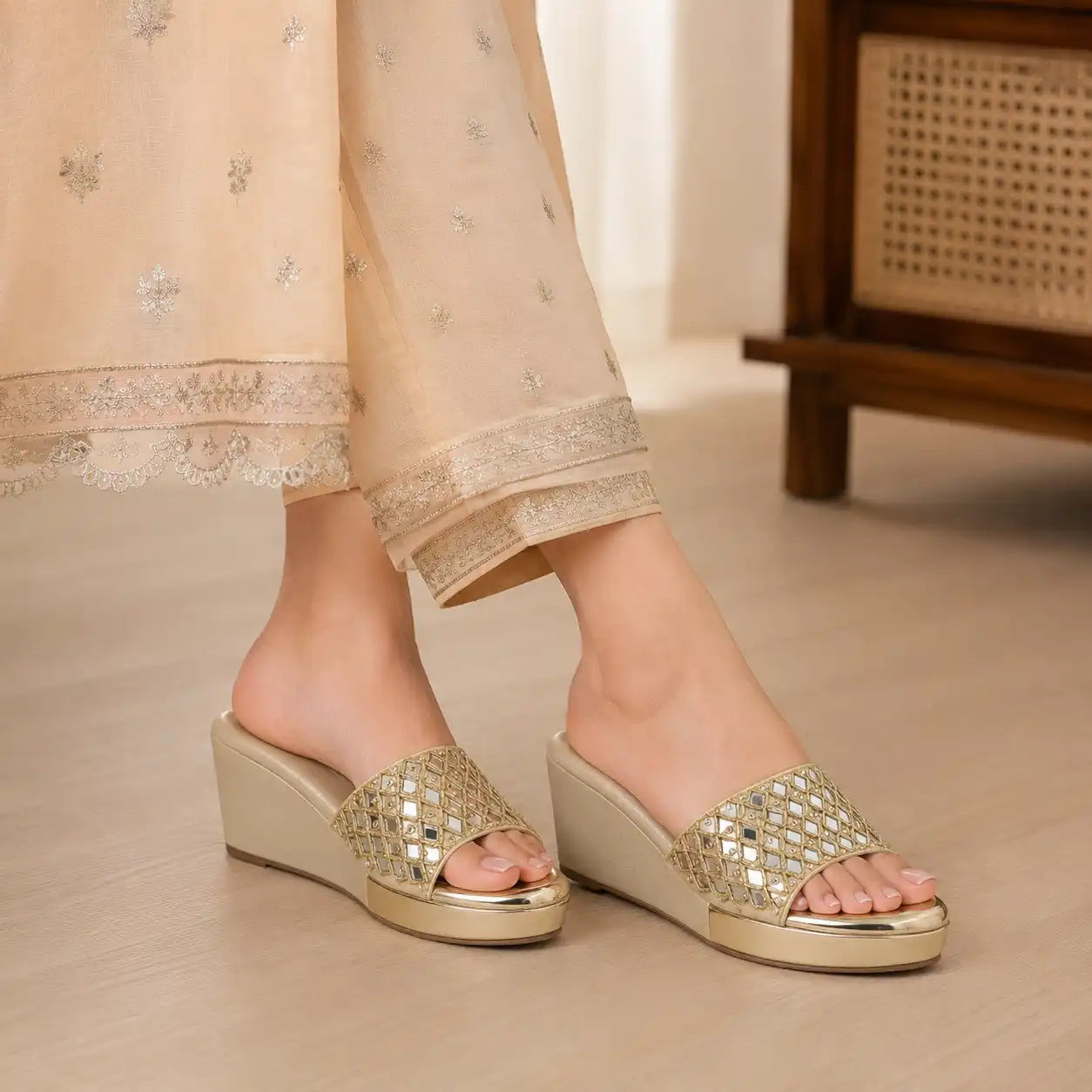 A person wearing beige embroidered pants and metallic gold wedge slide sandals with rhinestone-embellished straps, shown from the knees down in an indoor setting.