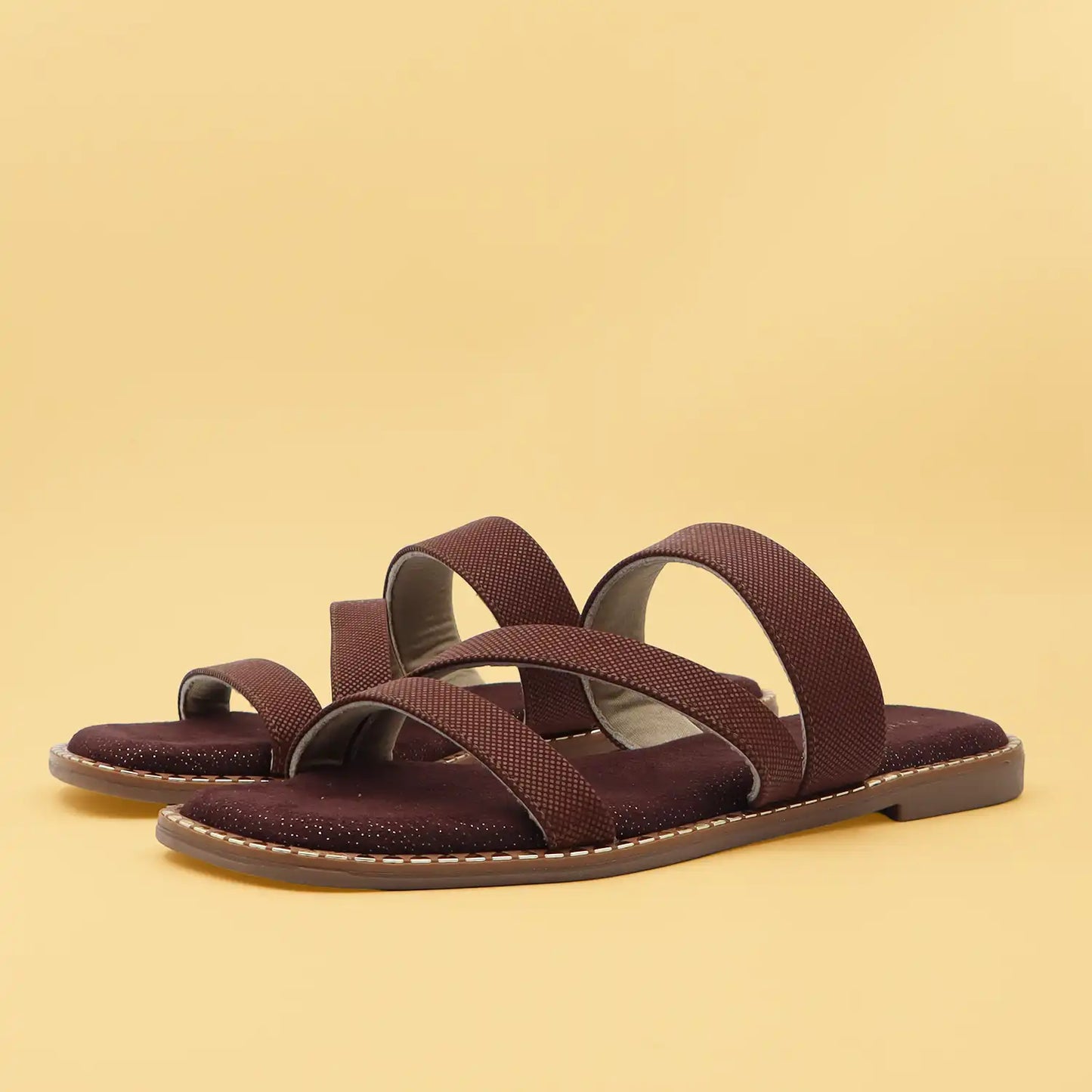 A pair of women's flat slide sandals in a deep burgundy-brown color, featuring textured straps and a glittery sole, set against a solid pale yellow background.