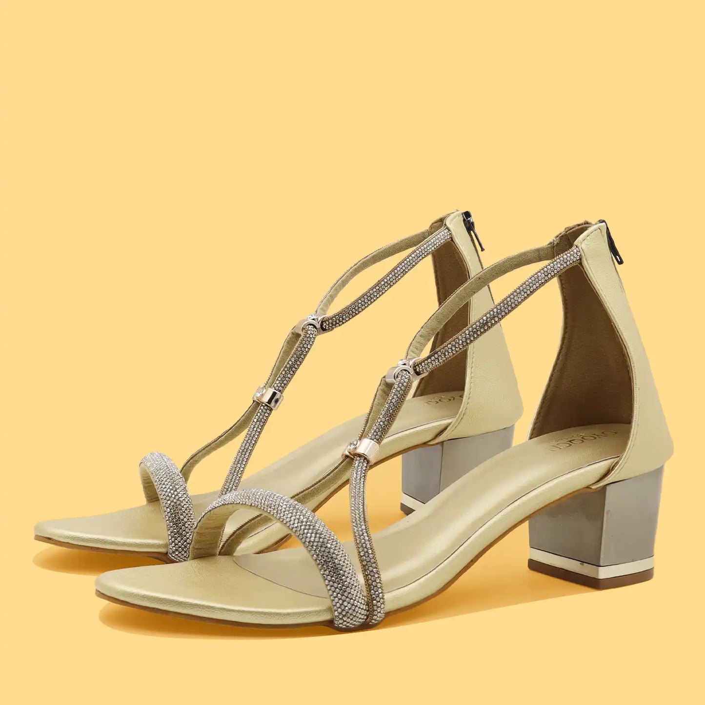 A pair of women's block-heel sandals in a light beige or champagne color, featuring shimmering silver rhinestone-embellished straps and a chunky silver block heel, set against a solid pale yellow background.