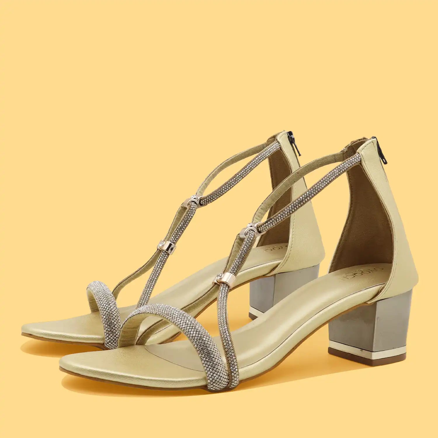 A pair of women's block-heel sandals in a light beige or champagne color, featuring shimmering silver rhinestone-embellished straps and a chunky silver block heel, set against a solid pale yellow background.